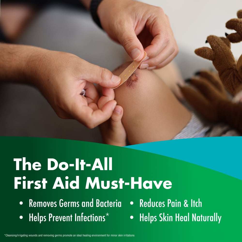 An adult places an adhesive bandage over a scrape on a child’s knee beside a stuffed animal. Text: “The Do-It-All First Aid Must-Have”; “Removes Germs and Bacteria”; “Helps Prevent Infections*”; “Reduces Pain & Itch”; “Helps Skin Heal Naturally”; “*Cleansing/irrigating wounds and removing germs promote an ideal healing environment for minor skin irritations”.